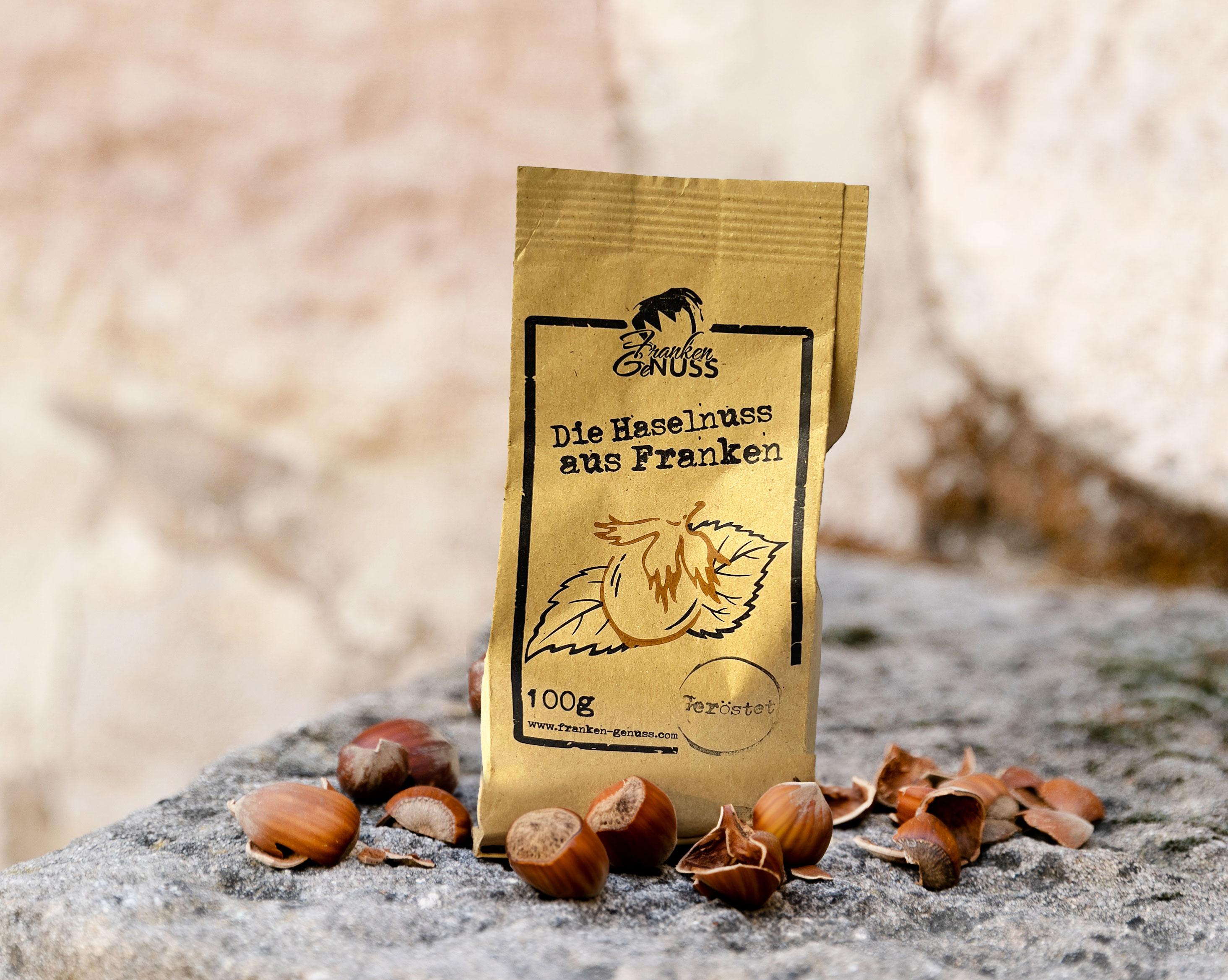 Eine rechteckige Verpackung steht auf einem grauen Untergrund, umgeben von ganzen Haselnüssen und Nussschalen. Eine rechteckige Verpackung steht auf einem grauen Untergrund, umgeben von ganzen Haselnüssen und Nussschalen.