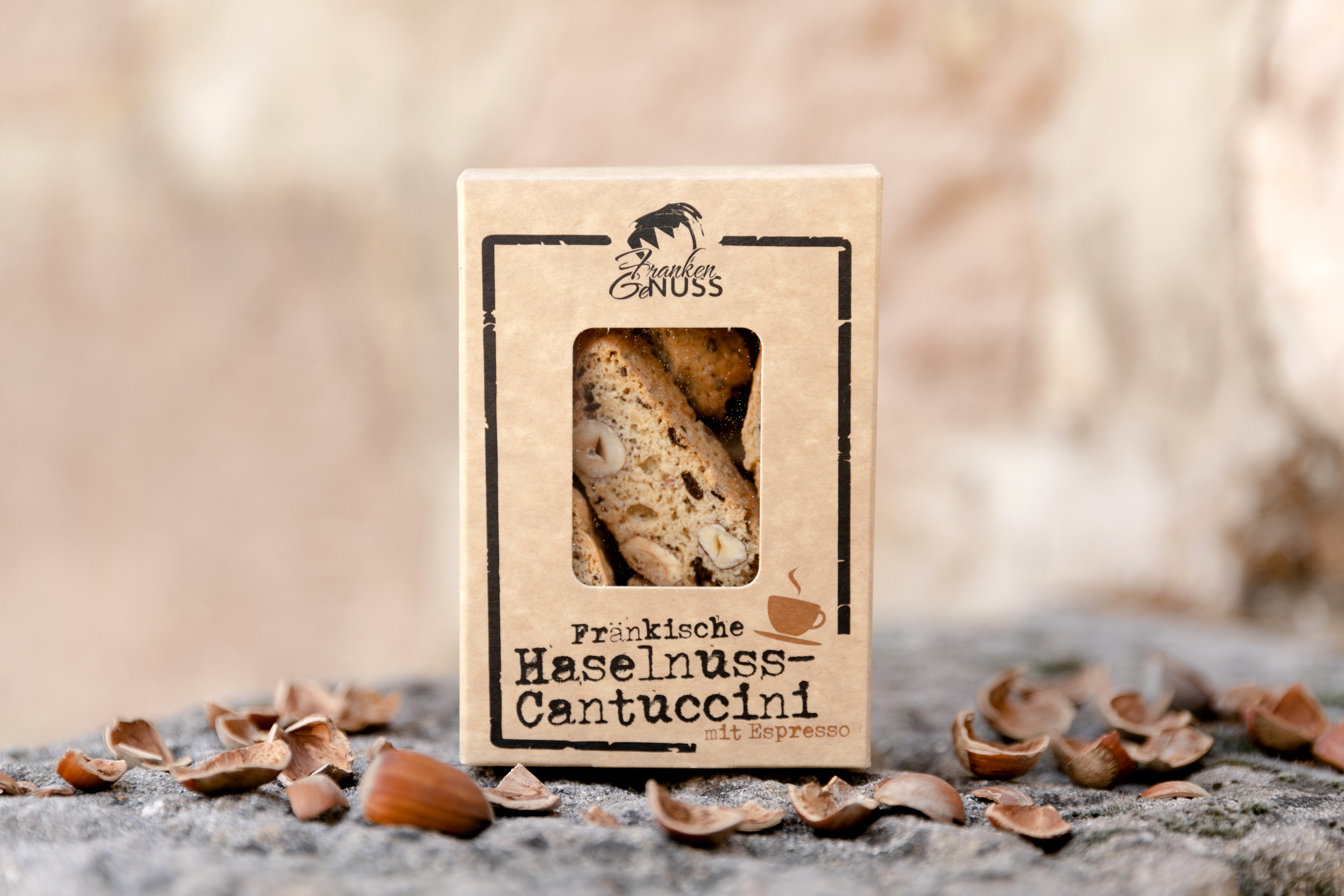 Knackige Haselnuss-Cantuccini mit Espresso in einer dekorativen Box, umgeben von ganzen Haselnüssen und Schalen auf Stein.