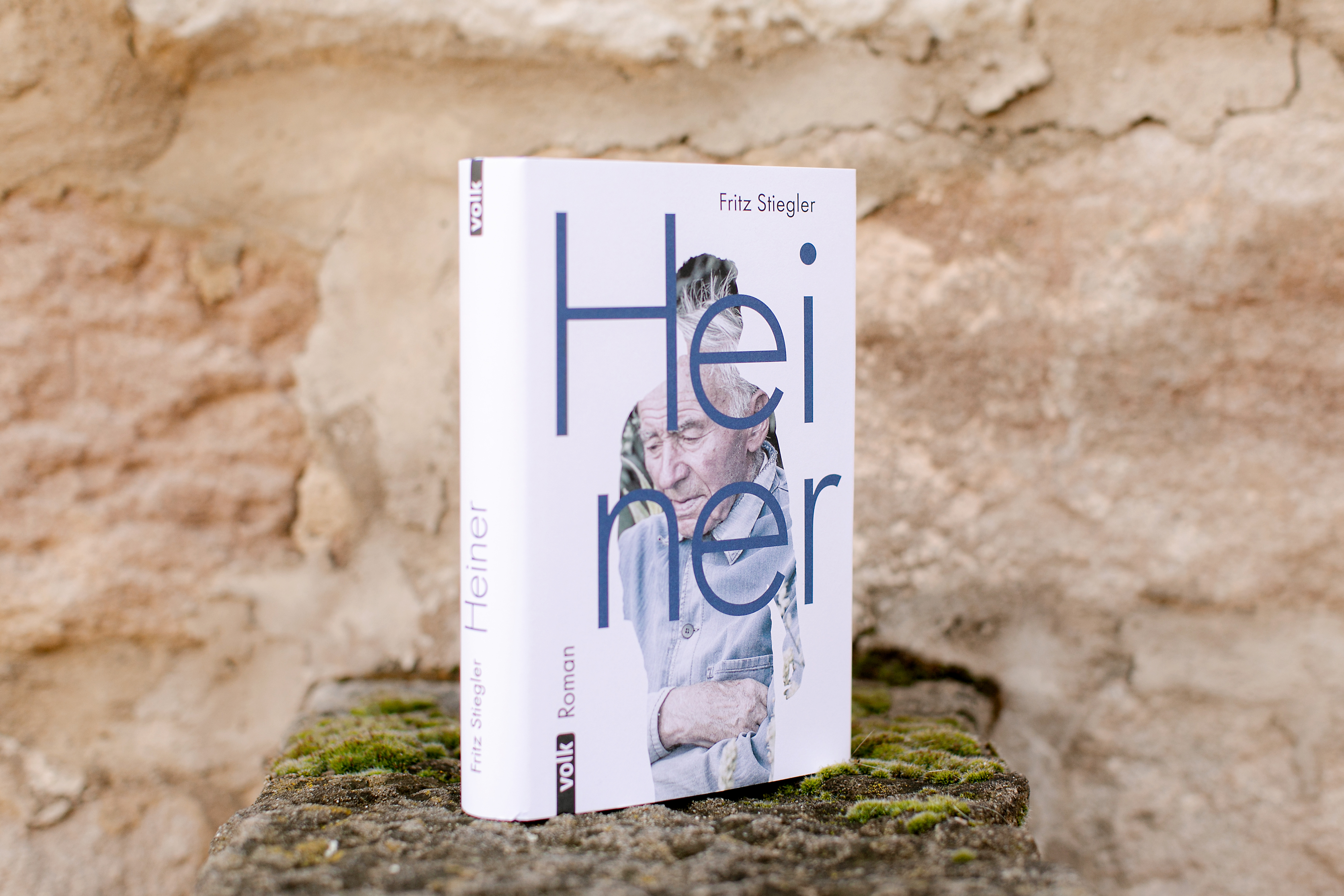 Das Buch Heiner steht auf einer rustikalen Steinmauer mit moosigem Untergrund vor einer hellen Wand.