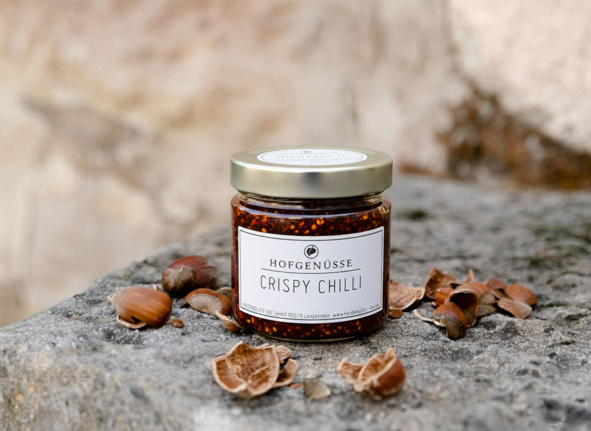 Ein Glas Crispy Chilli steht umgeben von Haselnüssen und Nussschalen auf einem steinernen Untergrund.