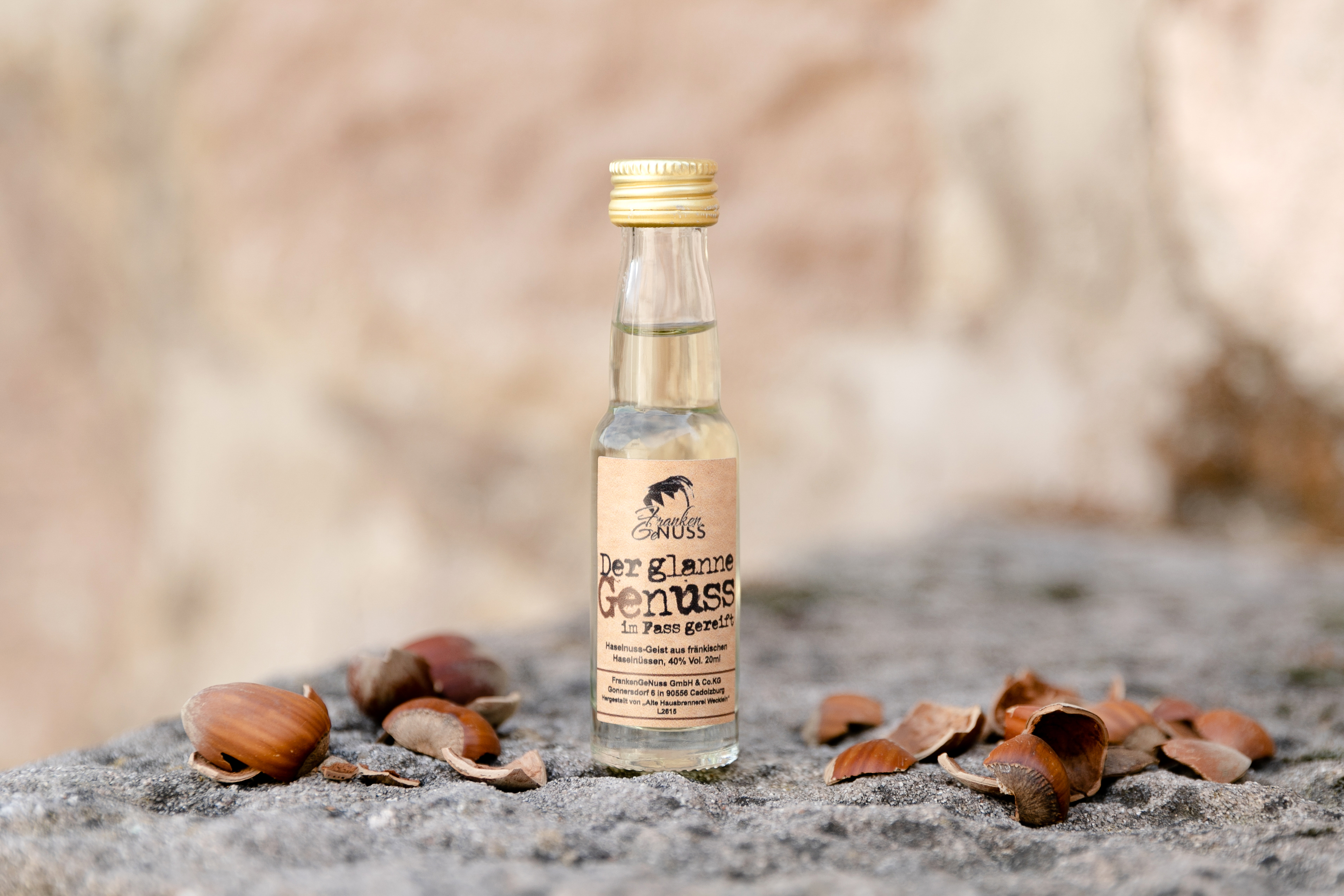 Eine kleine Glasflasche mit fassgereifter Haselnuss-Spirituose steht auf Stein, umgeben von frischen Haselnüssen und Nussschalen.