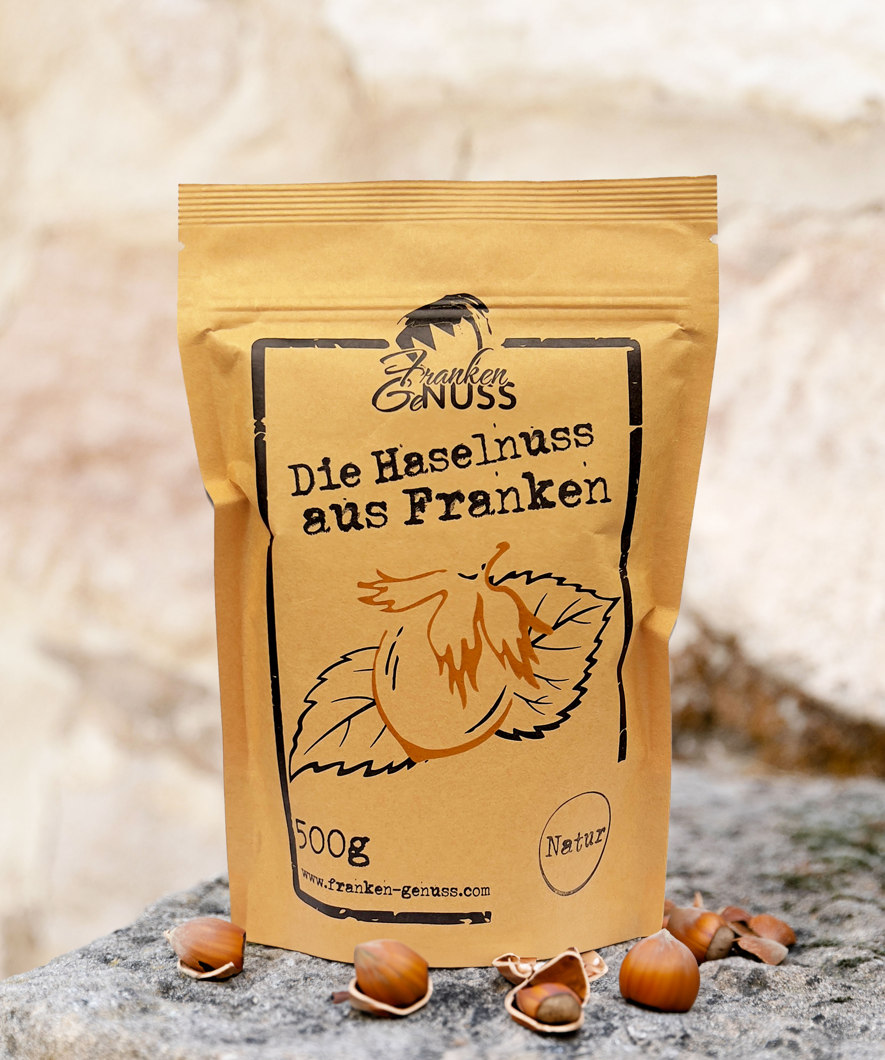 Eine rechteckige, hellbraune Verpackung mit Haselnuss-Grafik steht umgeben von echten Haselnüssen auf einer grauen Fläche.